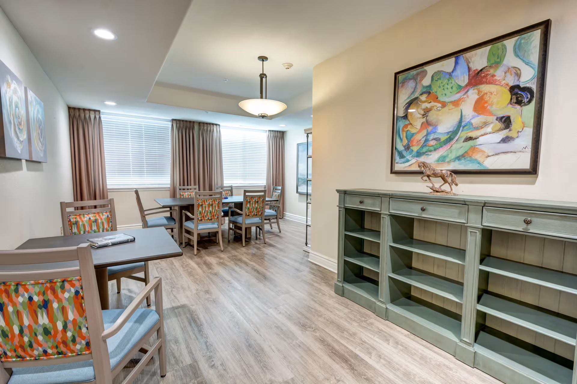 Bright communal dining area with tables, colorful patterned chairs, wood-look flooring and a green shelving unit beneath abstract artwork.