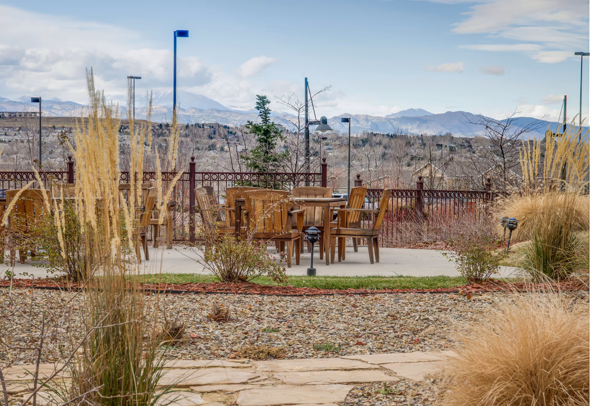 Outdoor patio area with wooden tables and chairs surrounded by ornamental grasses and shrubs, with a decorative metal fence and mountains visible in the background under a partly cloudy sky.