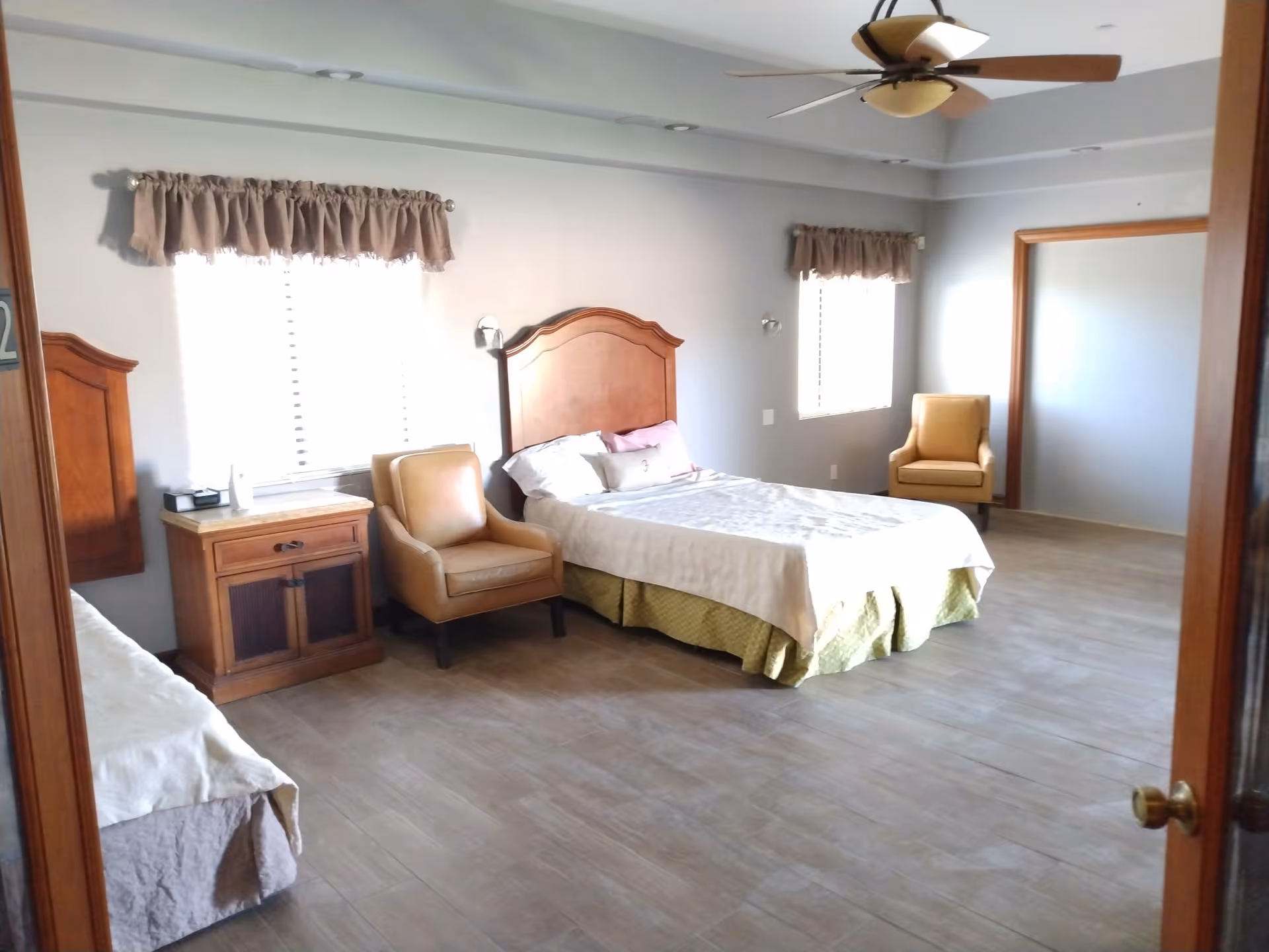 Spacious bedroom with two beds, wooden headboards, a nightstand, two armchairs, large windows with valances, a ceiling fan, and wood-look tile flooring.
