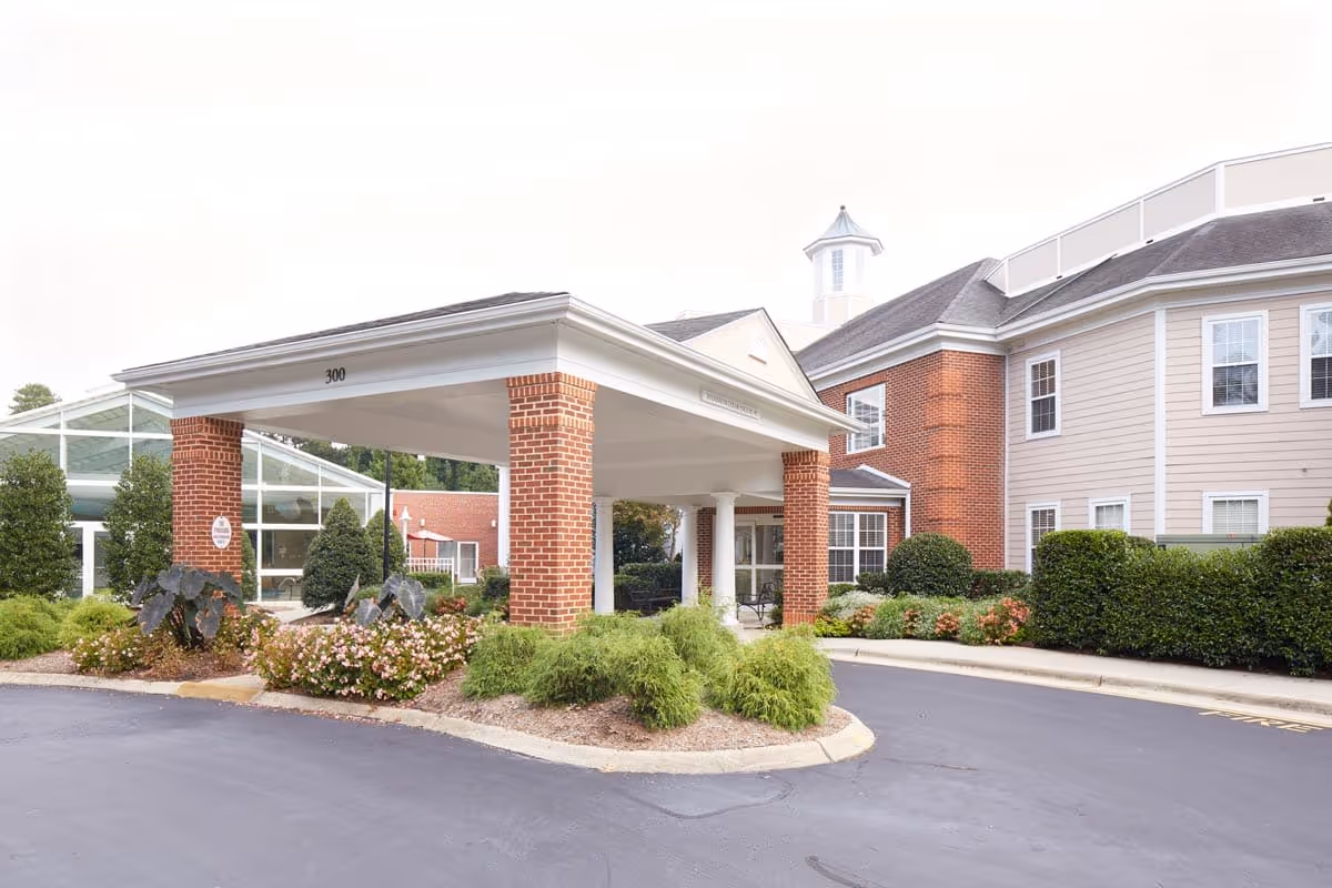 Entrance of a senior living facility named Woodland Terrace featuring a covered drop-off area with brick pillars, surrounded by well-maintained landscaping including bushes and flowers, and a multi-story building with beige siding and white-trimmed windows in the background.
