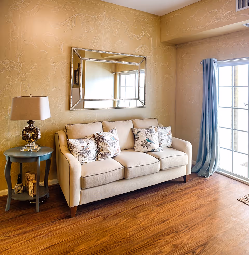 A cozy living room area featuring a beige three-seat sofa with four decorative pillows with bird and floral patterns. To the left of the sofa is a small round blue side table with a decorative lamp on top. Above the sofa hangs a large rectangular mirror with a reflective frame. The walls have a textured, patterned design in a warm beige tone. To the right, there is a large sliding glass door with light blue curtains, allowing natural light to brighten the room. The floor is a polished wood finish.