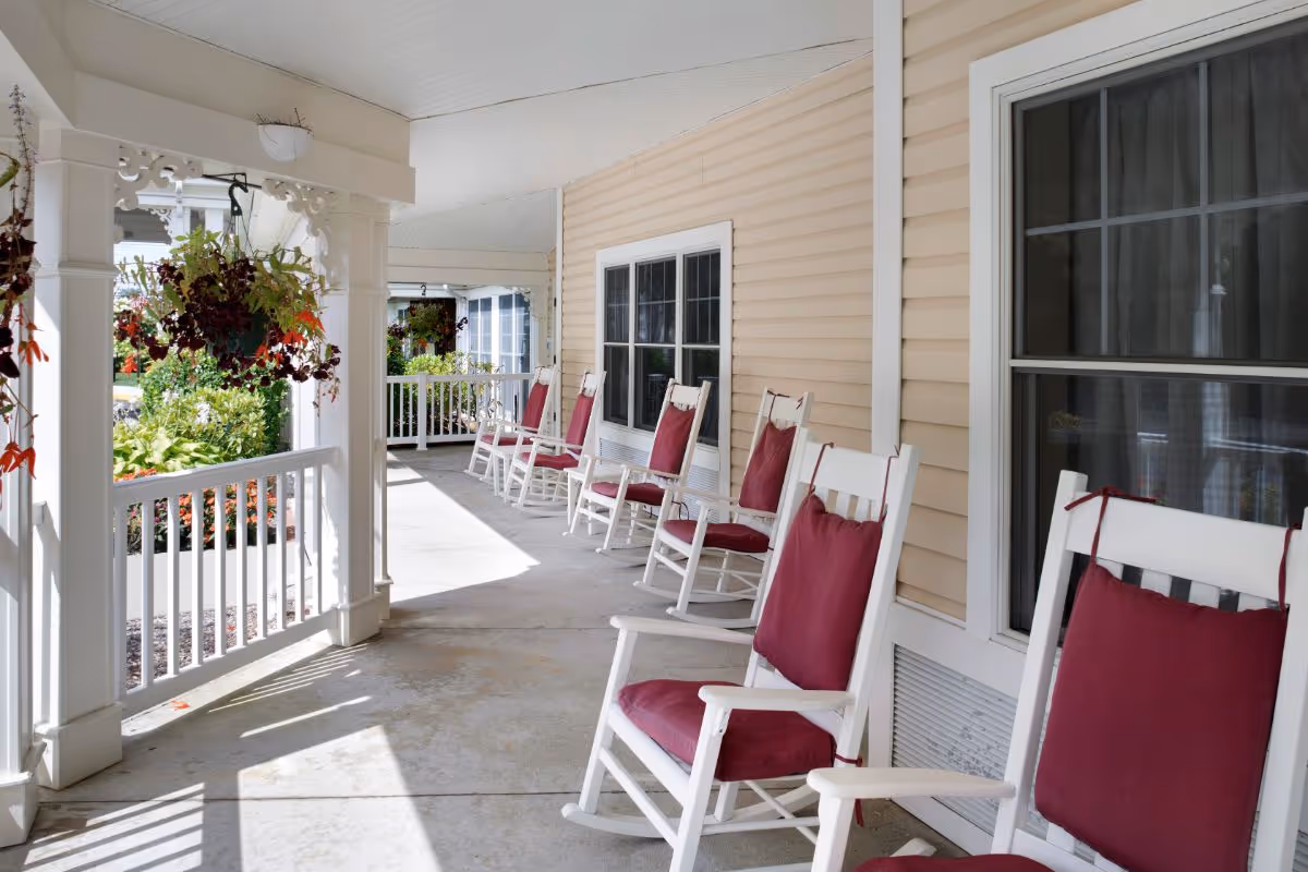 A covered porch area with a row of white rocking chairs each with a red cushion, beige siding on the building, hanging flower baskets, and a garden visible beyond the porch railing.