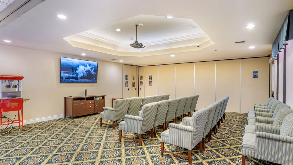 A small theater or presentation room with two rows of striped upholstered chairs facing a wall-mounted flat screen TV. There is a popcorn machine on the left side and a wooden cabinet below the TV. The room has patterned carpet, recessed lighting, and a ceiling-mounted projector.