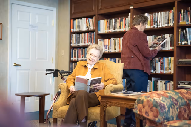 An older woman sits reading and smiling in a library-style common room while another person browses books on the shelves.