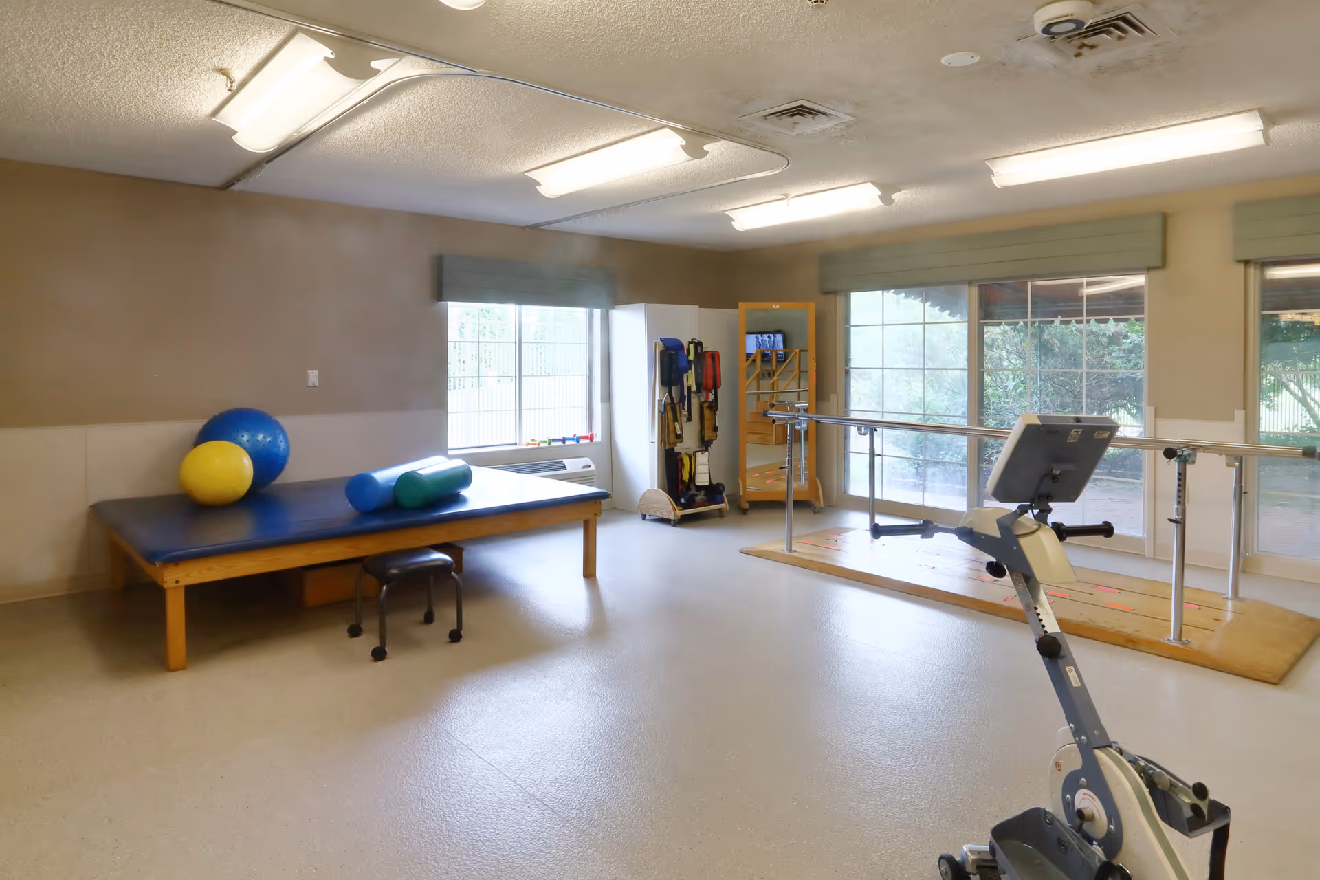 A physical therapy room with a padded treatment table holding exercise balls and foam rollers, a stool, parallel bars for walking exercises, a stationary exercise bike, and large windows letting in natural light.