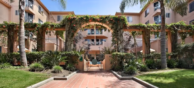 Outdoor view of a senior living facility entrance with a tiled walkway leading to a gated archway covered in greenery and flowers, surrounded by palm trees and landscaped gardens, with a multi-story building in the background under a clear blue sky.