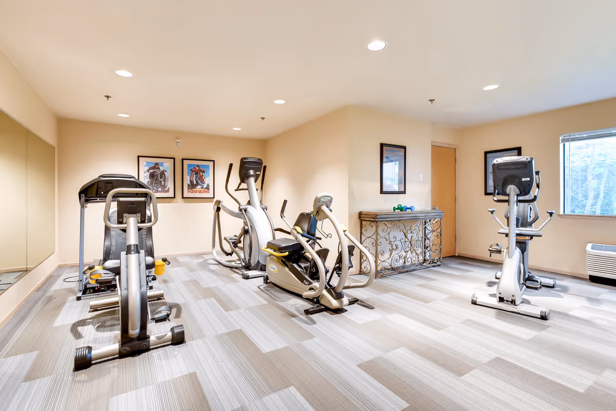 A small fitness room with exercise equipment including stationary bikes and elliptical machines. The room has beige walls, a patterned carpet floor, framed pictures on the walls, a large mirror on one side, and a window letting in natural light.