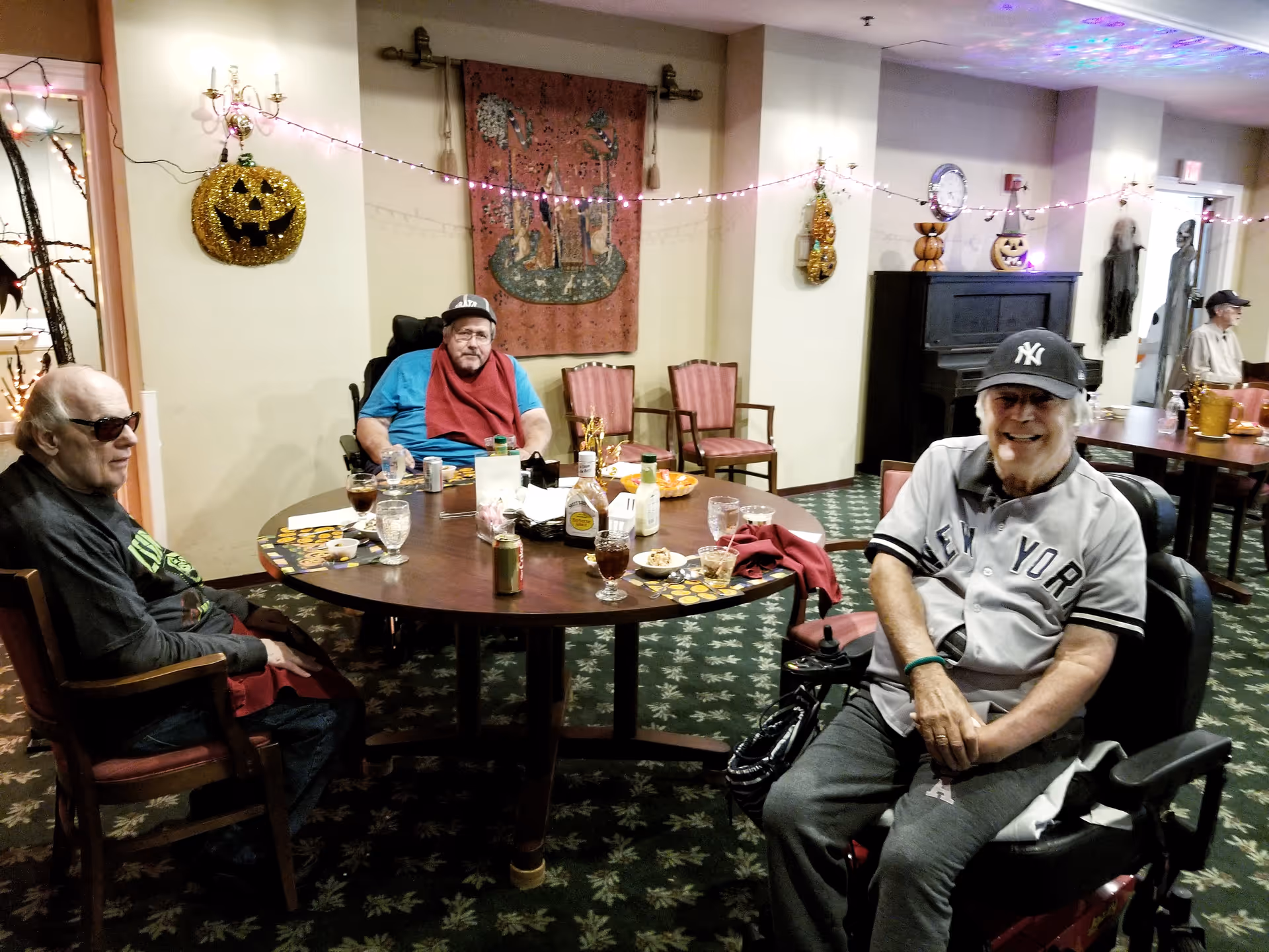 Three residents seated around a decorated communal dining table in a senior living facility lounge with Halloween decorations.