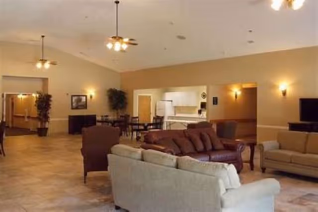 Spacious senior living common room with sofas, armchairs, a dining table and a small kitchen area under warm lighting.