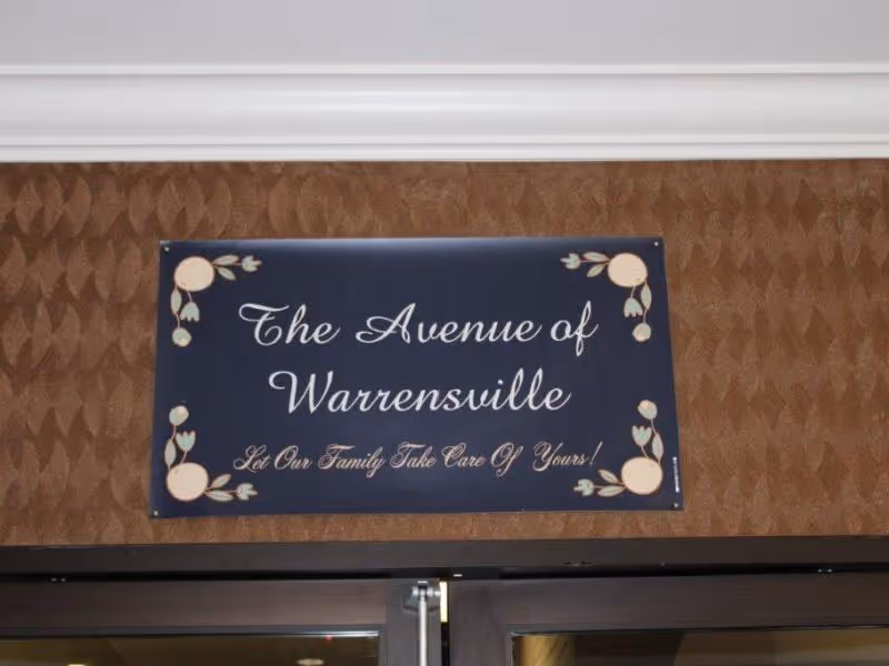 A dark blue sign with floral decorations on the corners mounted on a brown textured wall above a doorway. The sign reads 'The Avenue of Warrensville Let Our Family Take Care Of Yours!'