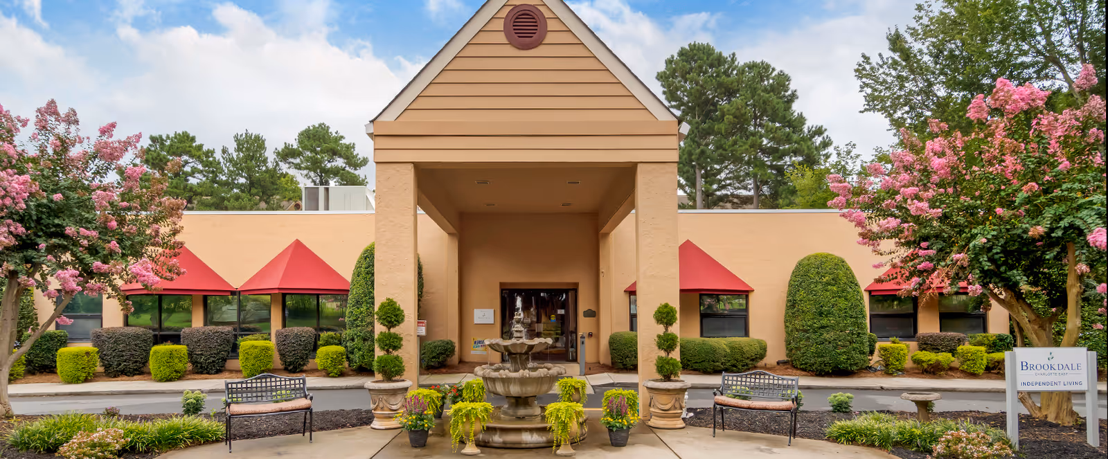 Front exterior view of Brookdale Charlotte East facility with a covered entrance, a central water fountain, two benches, potted plants, and landscaped bushes and trees with pink flowers on either side.