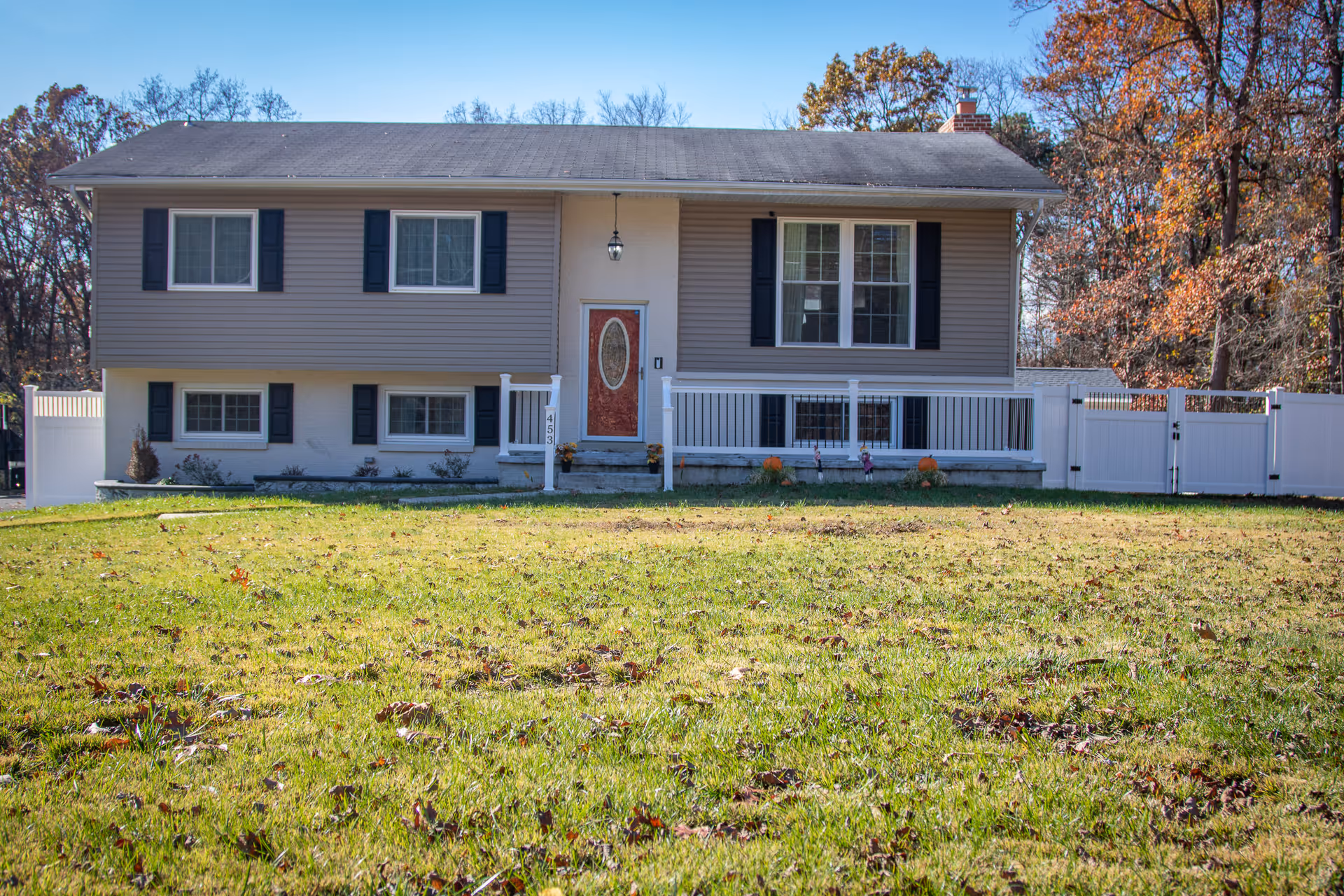 Front exterior view of a two-story house with gray siding and white trim, featuring a red front door with an oval window. The house has several windows with black shutters, a small porch with white railings, and a large grassy lawn in the foreground. Trees with autumn foliage are visible in the background.