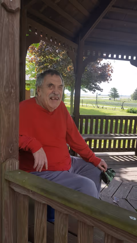 An elderly man wearing a red long-sleeve shirt and gray pants is sitting on a wooden bench inside a gazebo. The gazebo is made of wood with a slatted roof and railing. Behind him, there is a green lawn, trees with reddish leaves, and a fence in the background under a clear sky.