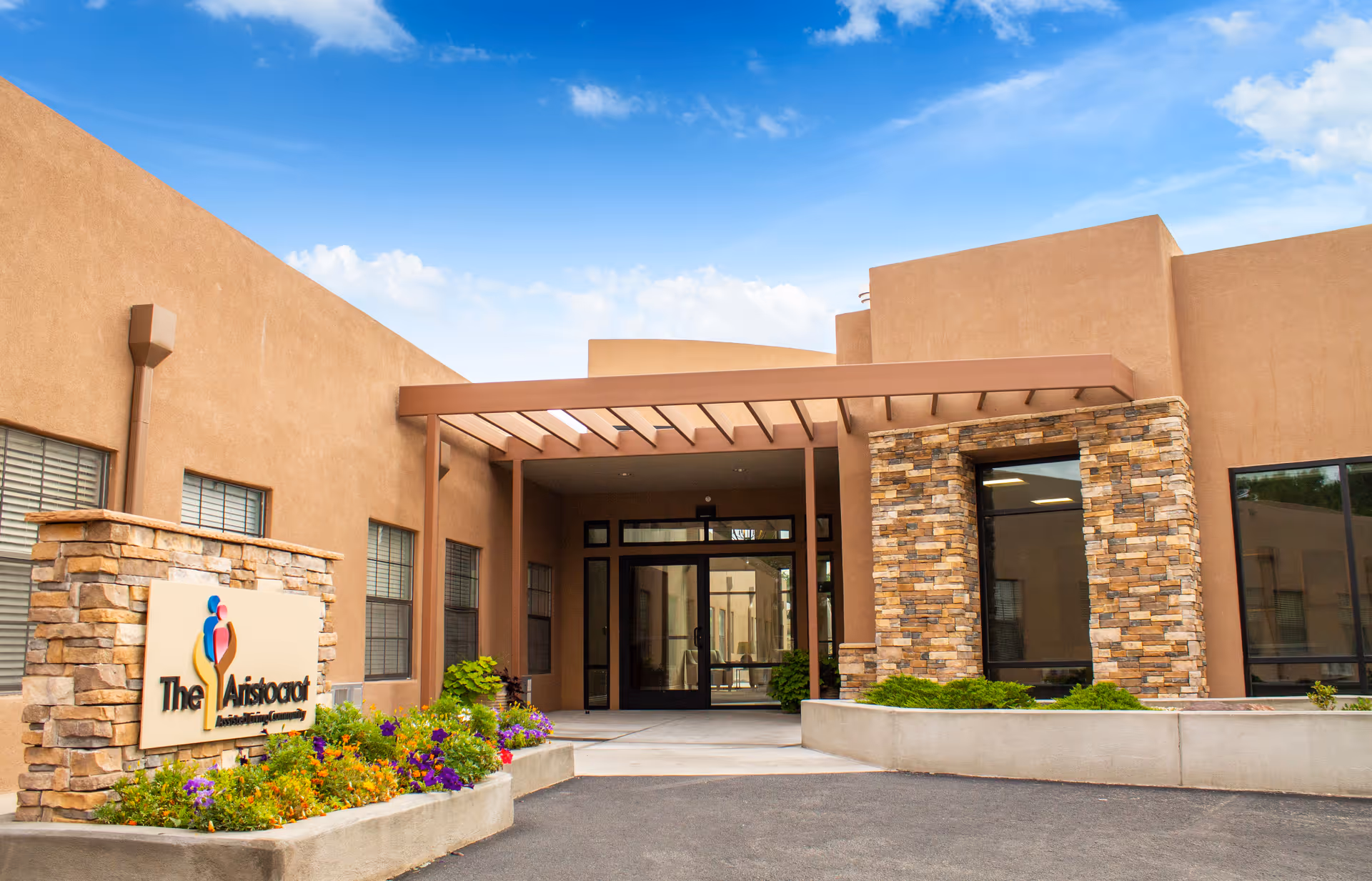 Front exterior entrance of an assisted living center with a stone sign reading 'The Aristocrat,' a covered entryway, and flowerbeds.