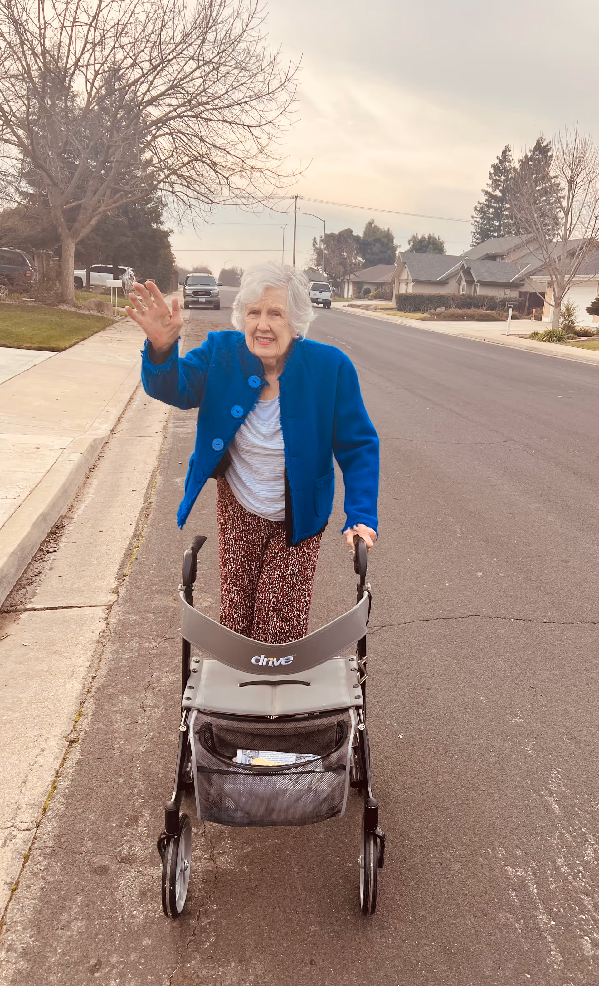 An elderly woman with white hair wearing a blue jacket and patterned pants is walking on a suburban street using a gray walker. She is smiling and waving with her left hand. The street is lined with houses, leafless trees, and parked cars under a cloudy sky.