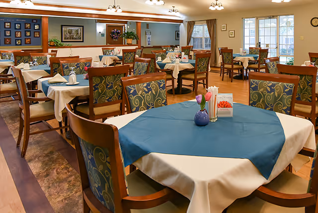 A dining room with multiple tables covered in white tablecloths and blue table runners. Each table is surrounded by wooden chairs with patterned upholstery. The room is well-lit with ceiling lights and has windows with curtains, framed pictures on the walls, and a clock. A small vase with flowers and a menu card are placed on the closest table.