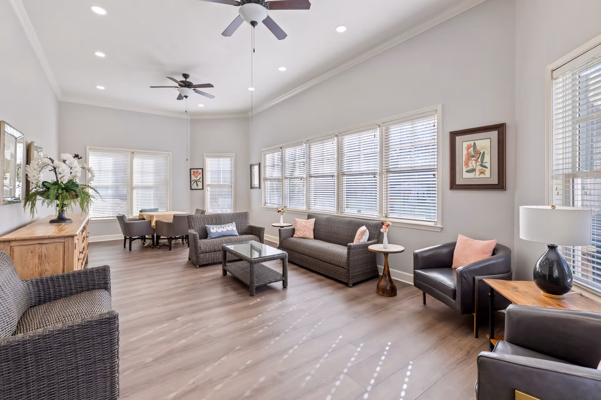A bright and spacious living room area with large windows covered by white blinds allowing natural light to fill the room. The room features wicker and leather seating including two sofas, two armchairs, and a wicker chair. There is a glass coffee table in the center, side tables with small flower vases, a wooden sideboard with a large floral arrangement, and framed botanical artwork on the walls. The floor is light wood, and the ceiling has recessed lighting and two ceiling fans.