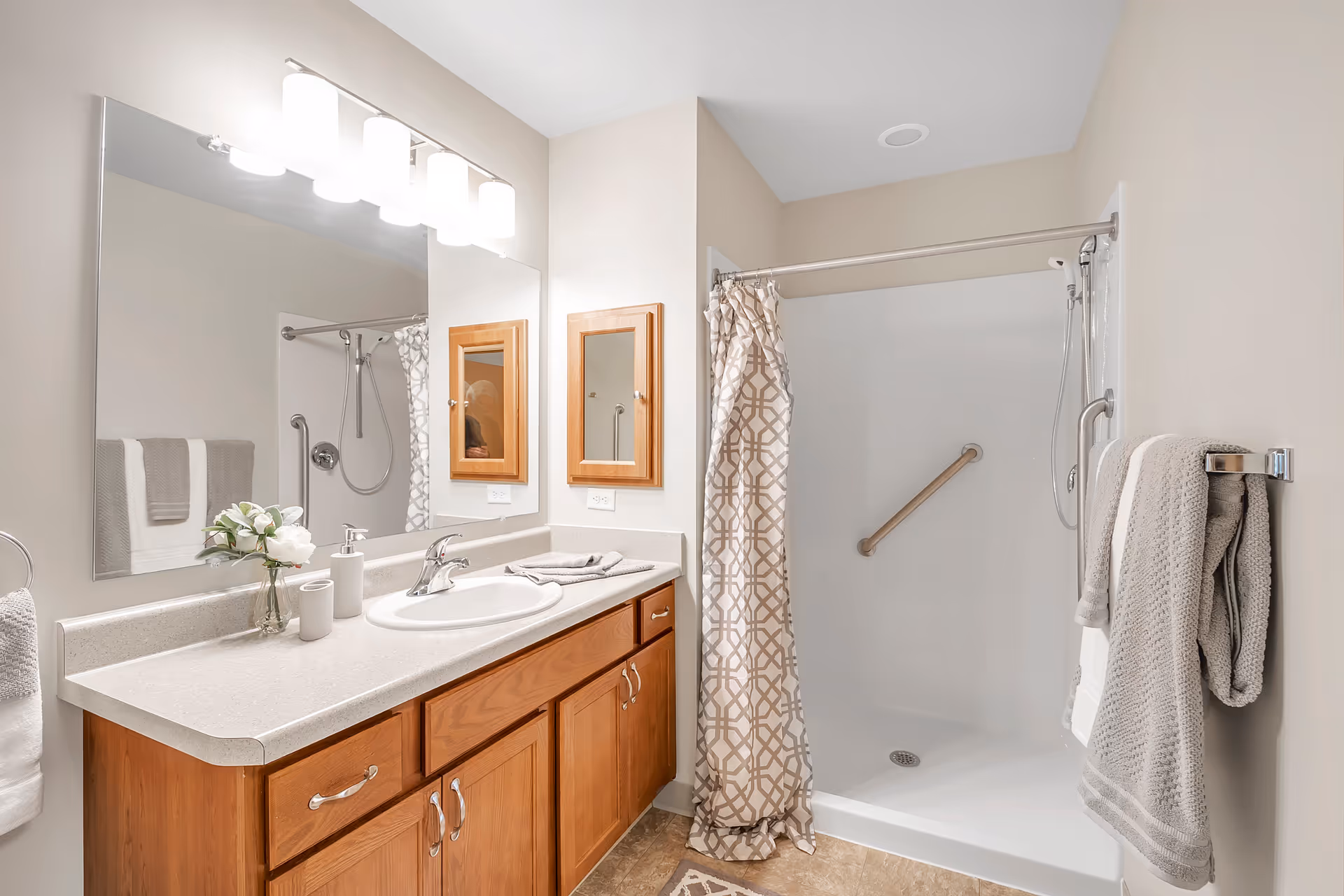 A clean and well-lit bathroom featuring a wooden vanity with a white countertop and sink. Above the vanity is a large mirror with four modern light fixtures. To the right is a walk-in shower with a patterned beige and white shower curtain, a grab bar, and a handheld showerhead. Towels are neatly hung on towel racks on both sides of the bathroom. The walls are painted a light beige color.