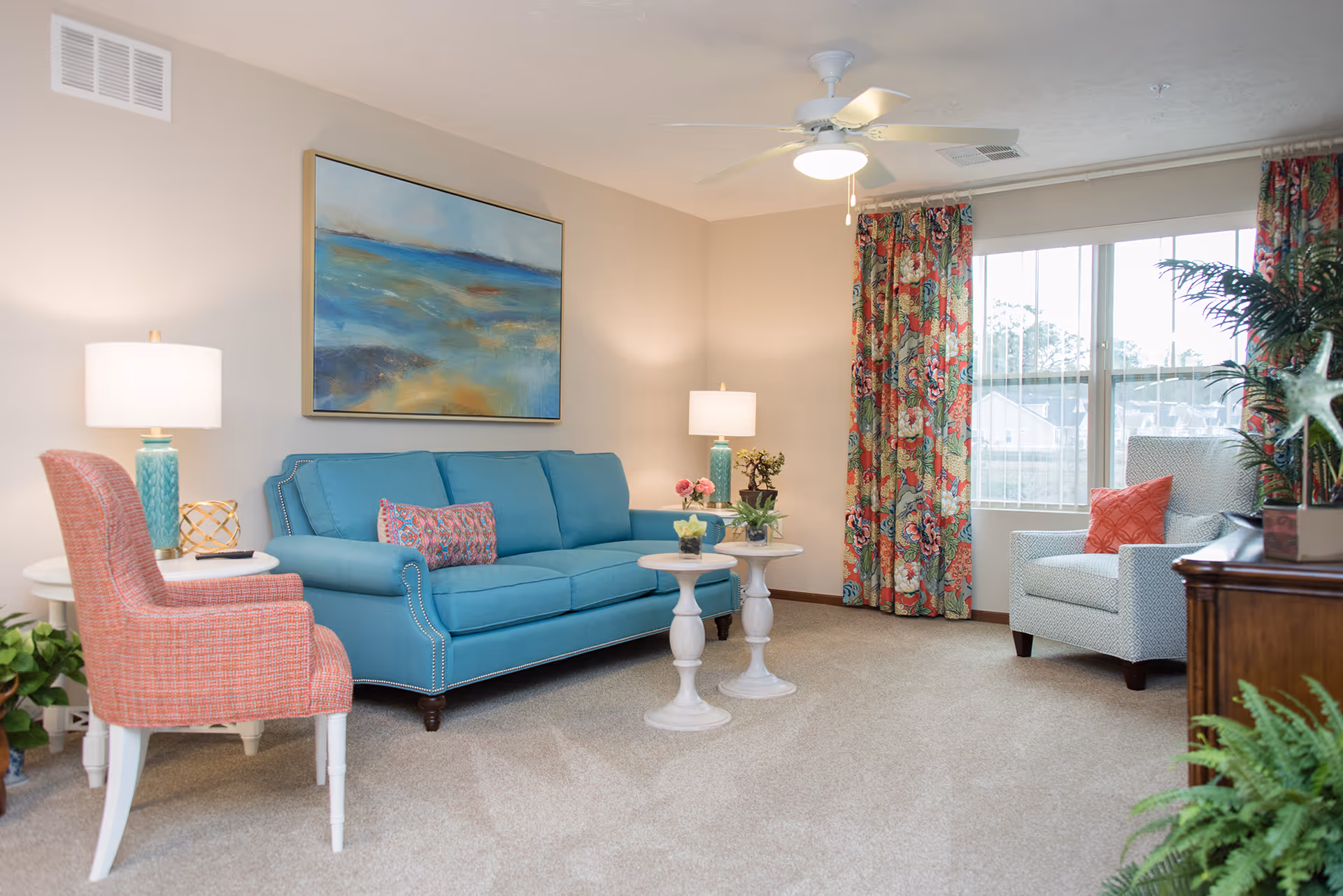 A bright and cozy living room with a blue sofa adorned with a colorful pillow, a coral armchair, two white round side tables with small plants, a large window with floral curtains, a ceiling fan with light, and a large abstract painting on the wall.