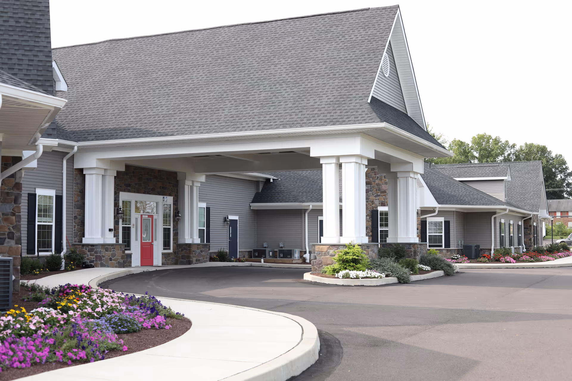 Exterior view of Bristol House Memory Care facility showing a covered entrance with white columns, stone and gray siding, a red door, and landscaped flower beds along the driveway.
