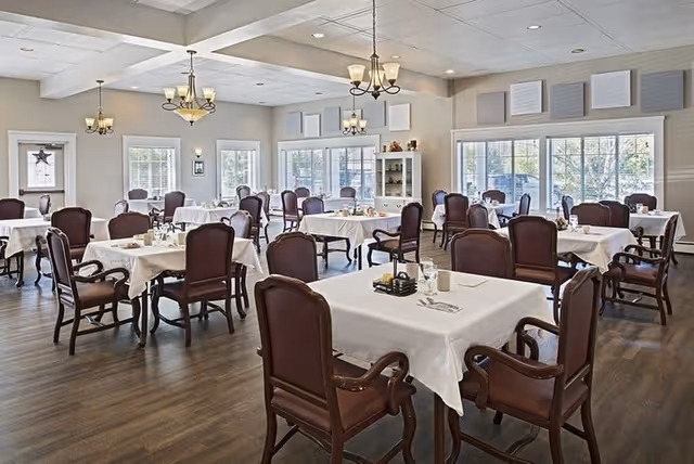 A bright and spacious dining room in a senior living facility with multiple tables covered in white tablecloths, each surrounded by wooden chairs with cushioned seats and backs. The room features large windows allowing natural light to fill the space, chandeliers hanging from the ceiling, and a cabinet with decorative items against the far wall.