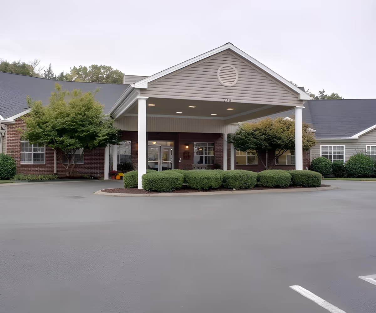 Front exterior view of Lincoln Manor senior living facility showing a covered entrance with white pillars, neatly trimmed bushes, trees, and a paved driveway.