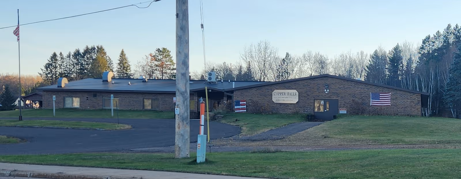 Exterior view of Copper Falls Assisted Living, a single-story brick building with an American flag on the right side and a flagpole with a flag at half-mast on the left side. The building is surrounded by grass and trees in the background under a clear sky.