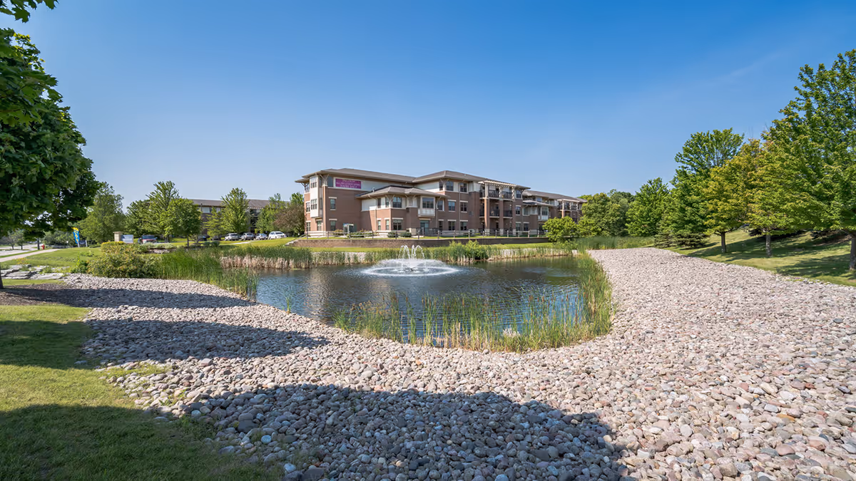 A three-story senior living building beside a landscaped pond with a central fountain and surrounding trees.