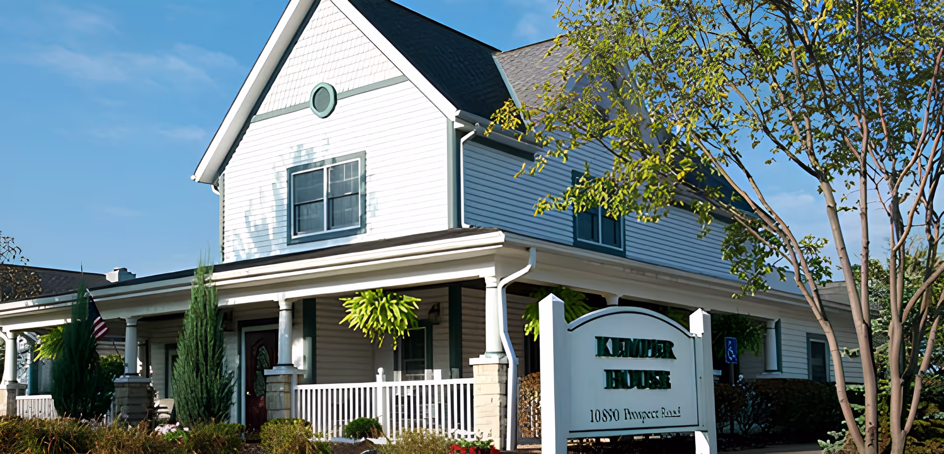 Exterior view of Kemper House Strongsville, a two-story white building with green trim, a covered porch with columns, hanging plants, and surrounding greenery under a clear blue sky.
