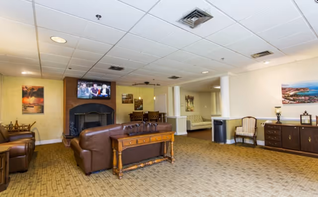A spacious living room area in a senior living facility with brown leather sofas, a wooden table, a fireplace with a TV mounted above it, and various paintings on the walls. The room has beige carpet flooring, a drop ceiling with recessed lighting, and a small dining area with chairs in the background.