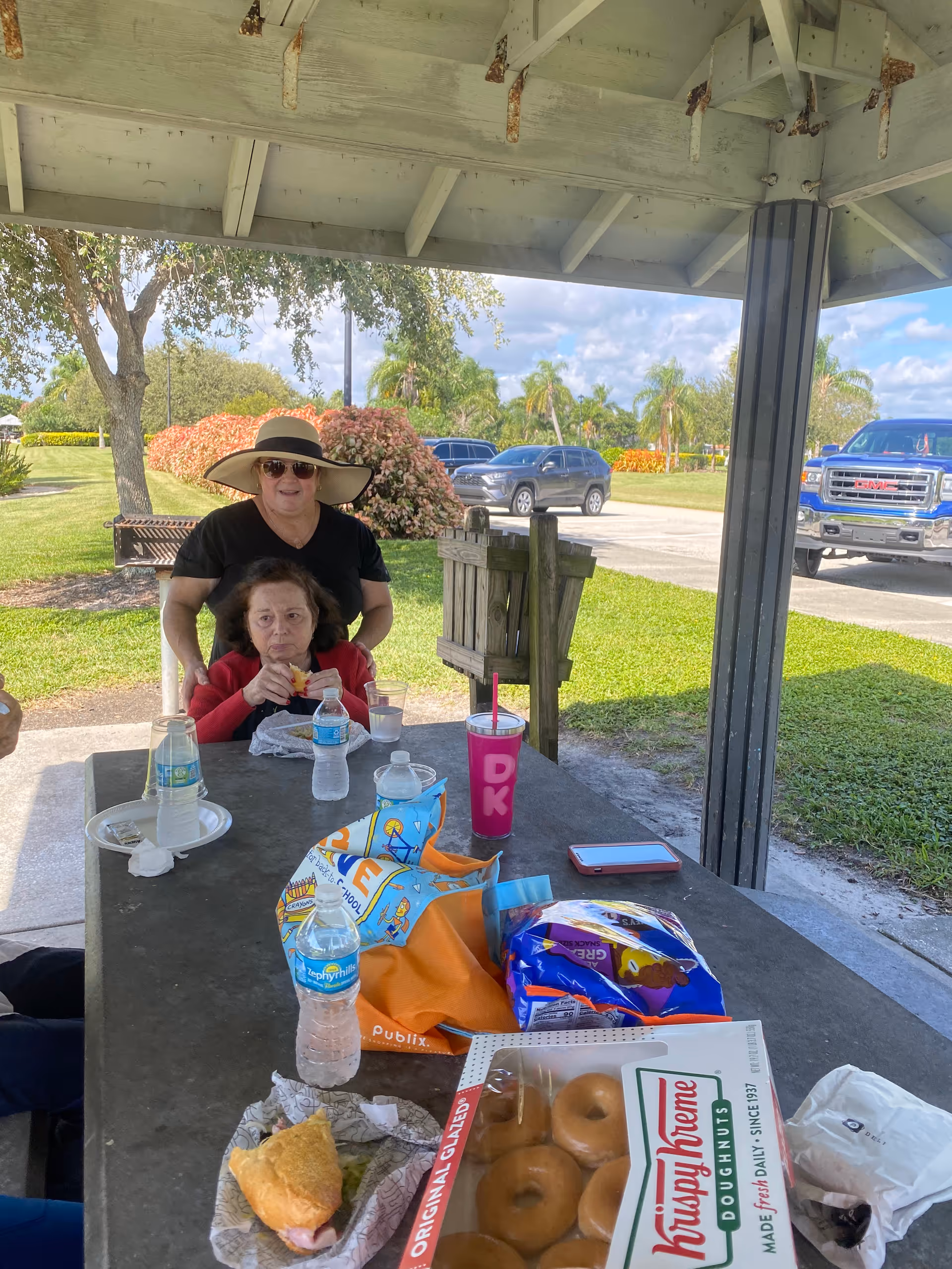 Two women sit at a picnic table under a pavilion eating snacks, with bottled water, a box of doughnuts, drinks, and bags on the table and cars and landscaping in the background.