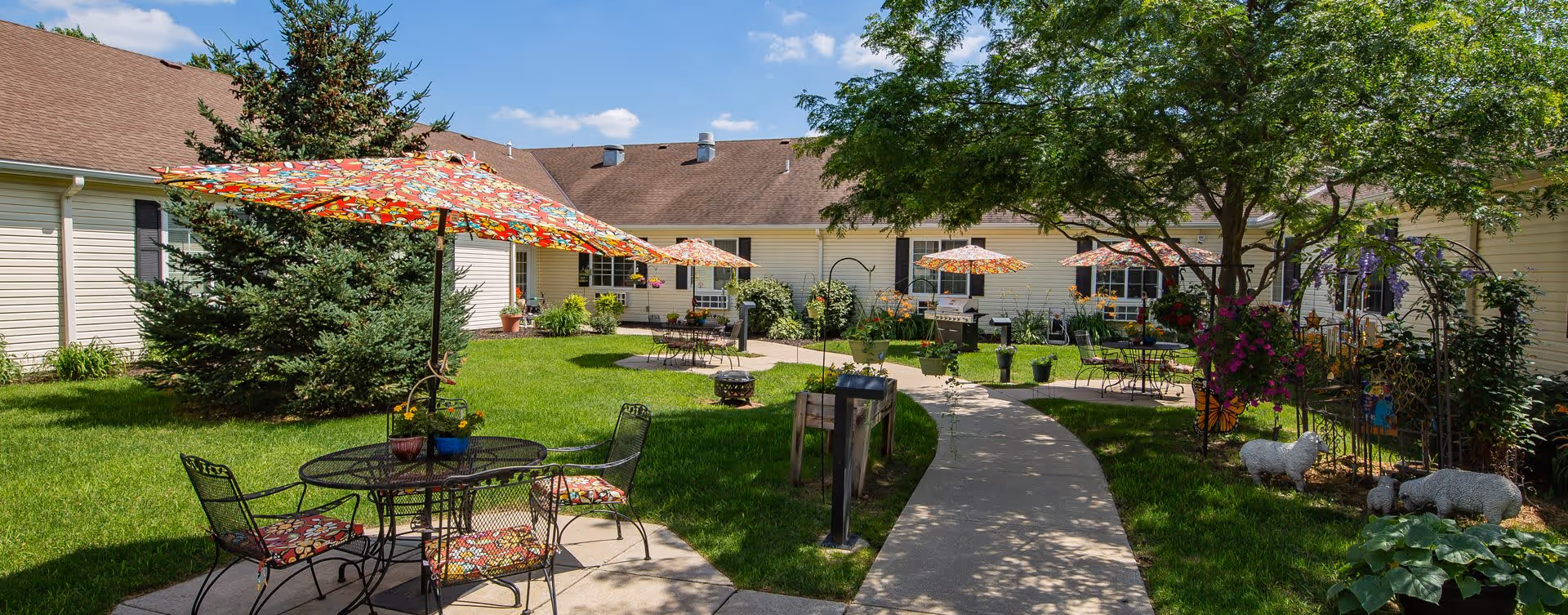 Outdoor courtyard area with green grass, colorful floral umbrellas shading metal tables and chairs, a paved walkway, various plants and flowers, and decorative garden sculptures including sheep figures. The courtyard is surrounded by a single-story building with beige siding and brown roof under a blue sky with some clouds.