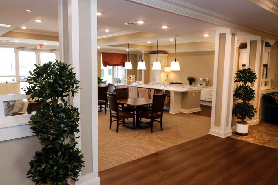 Interior view of a senior living facility showing a dining area with a round table and four chairs, a kitchen island with pendant lights hanging above, and decorative plants placed near white columns. The space is well-lit with recessed ceiling lights and has a warm, inviting atmosphere.