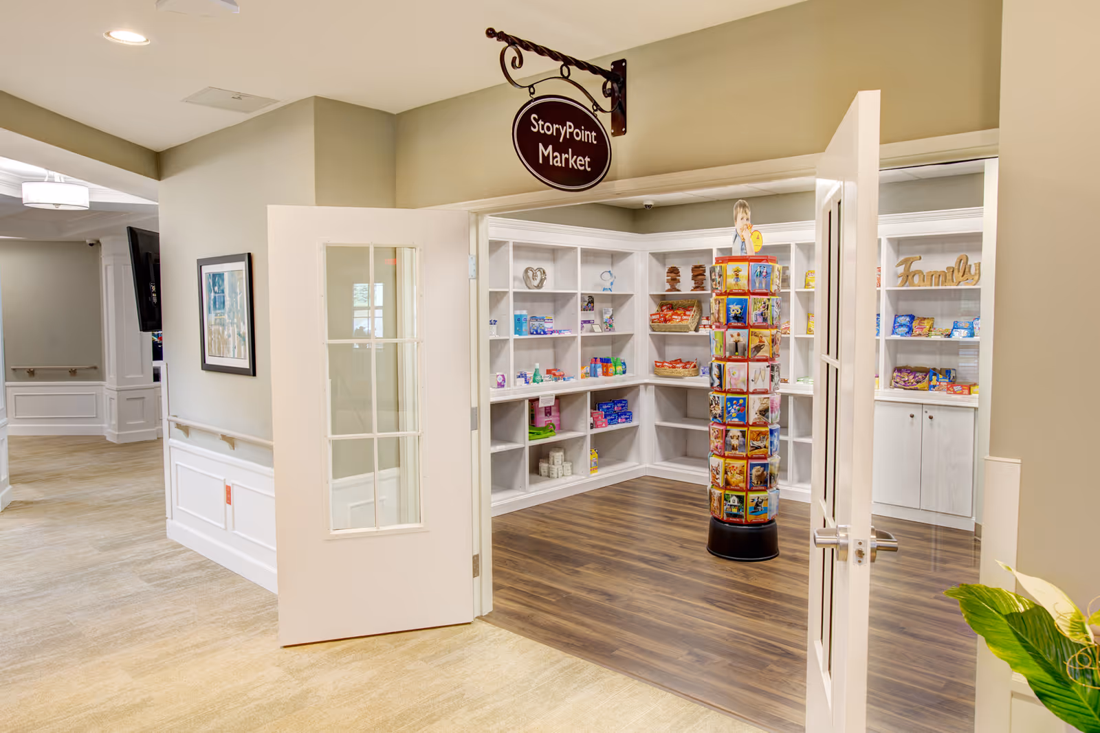 Interior view of a small market room named StoryPoint Market inside a senior living facility. The room has white shelves stocked with various snacks and items, a rotating display rack with greeting cards or similar products, and double doors open to the hallway with beige walls and light wood flooring.