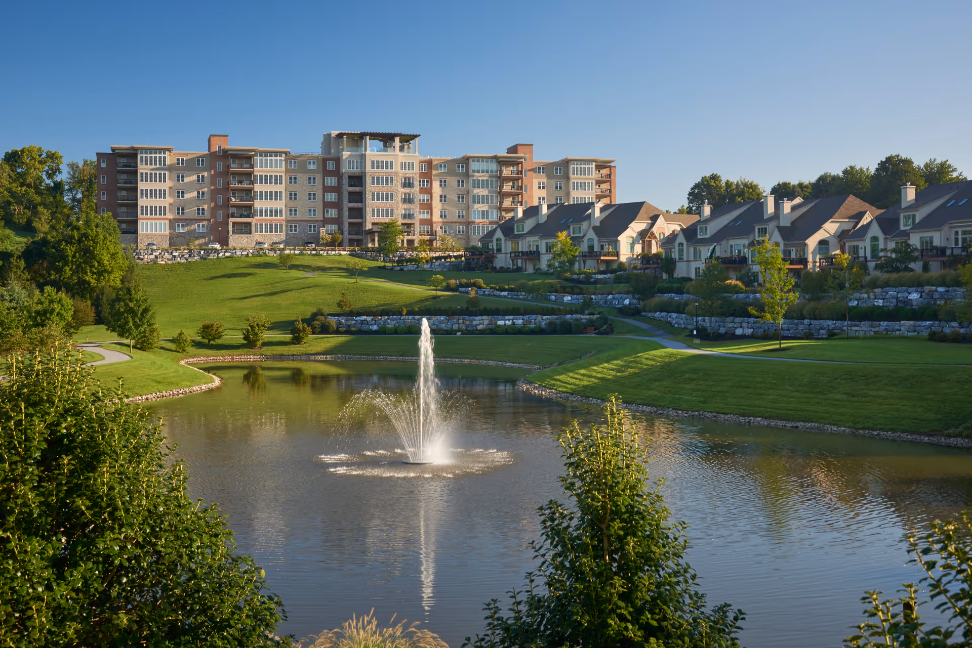 A scenic view of The Vistas at Willow Valley Communities featuring a large pond with a central water fountain, surrounded by well-maintained green lawns and trees. In the background, there is a multi-story residential building and a row of smaller townhouse-style homes under a clear blue sky.