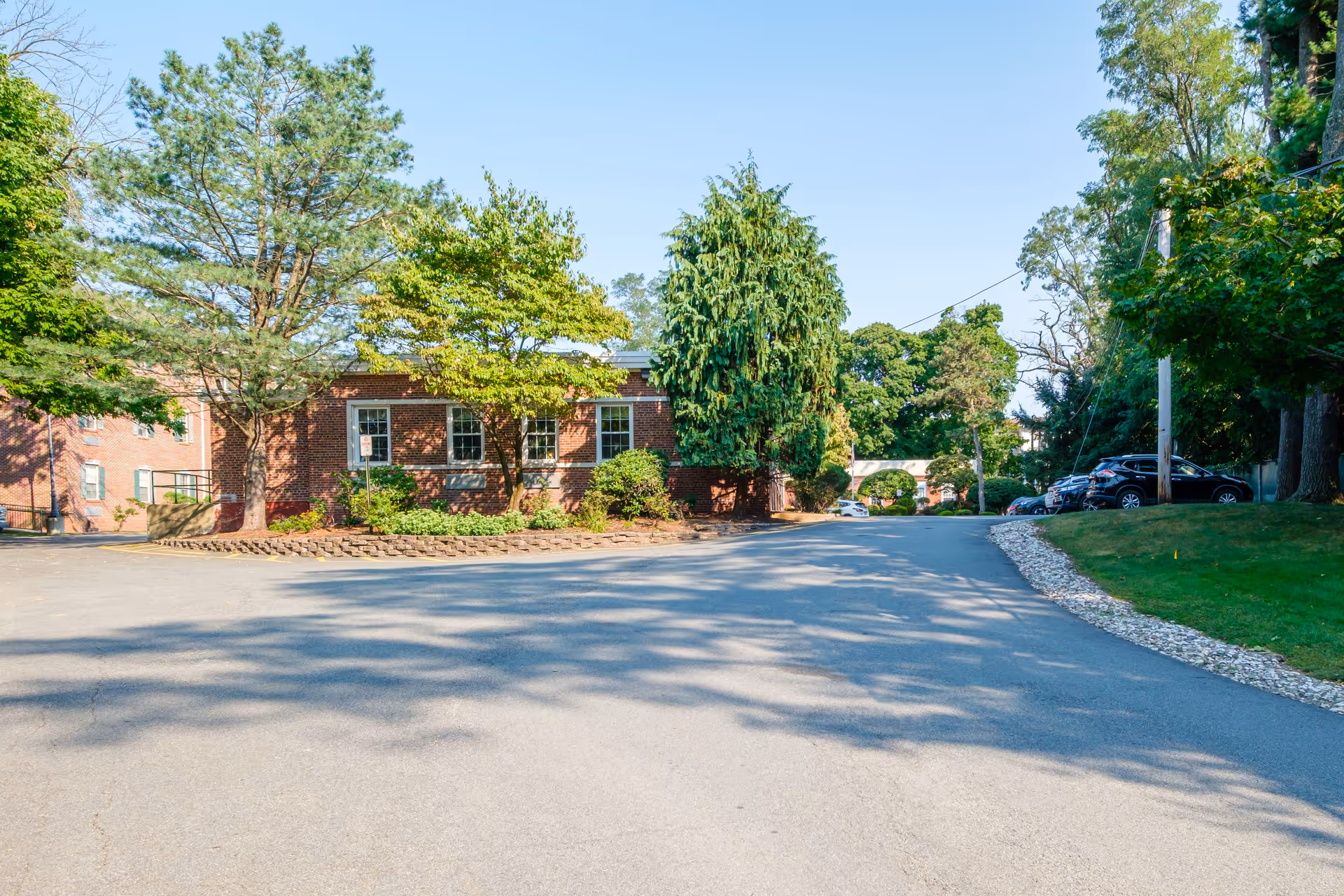 Paved driveway leading past a low brick building surrounded by trees and parked cars under a clear blue sky.