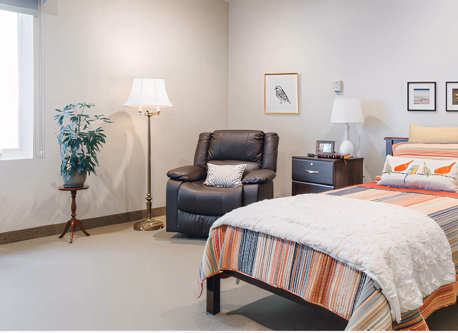 A bright furnished bedroom with a bed, recliner chair, nightstand, floor lamp, and potted plant.
