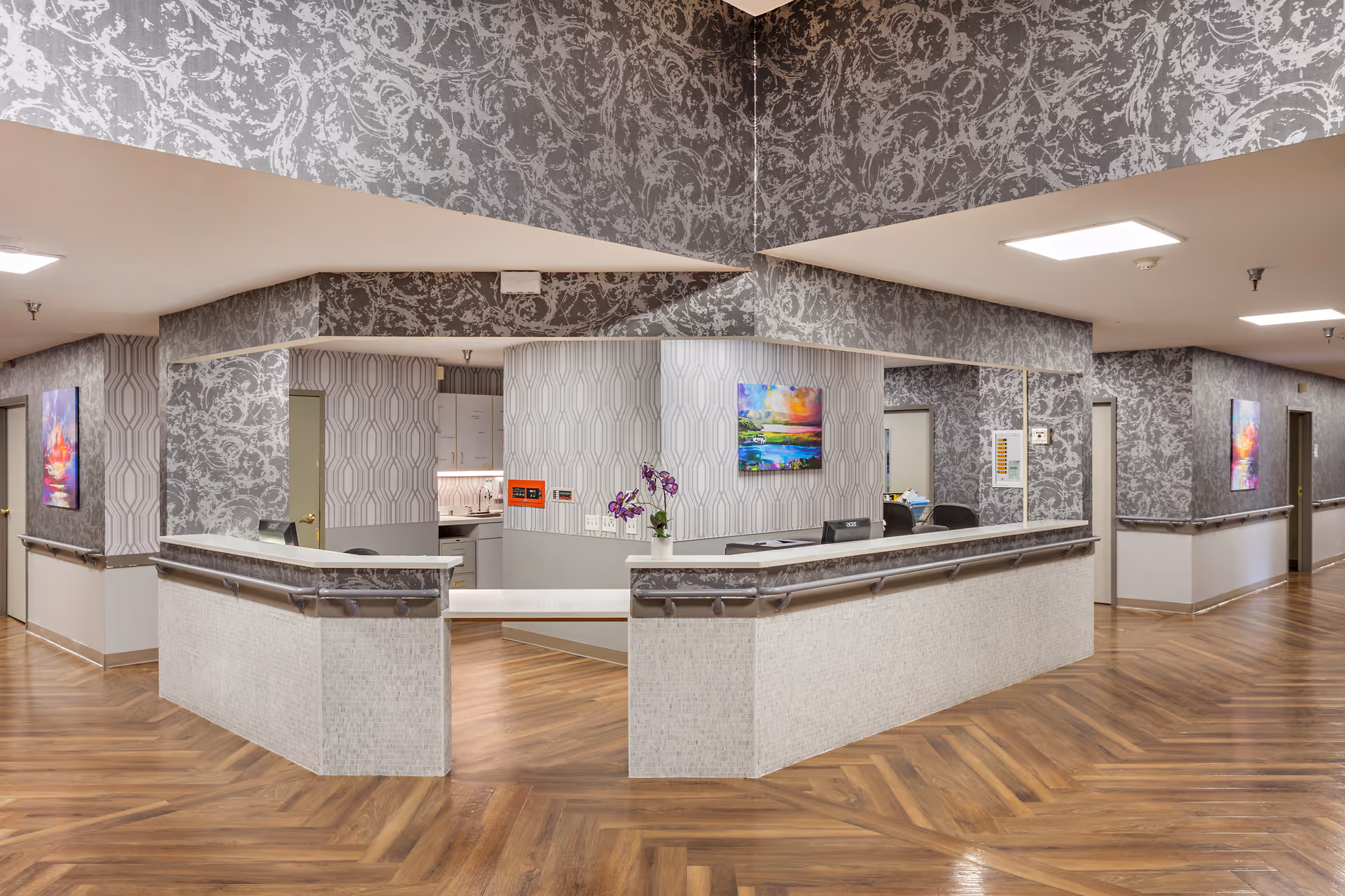 A modern reception desk area in a senior living facility with patterned wallpaper, wood flooring, and colorful artwork on the walls. The desk has a computer monitor and a small vase with purple flowers. The hallway extends on both sides with doors and handrails along the walls.
