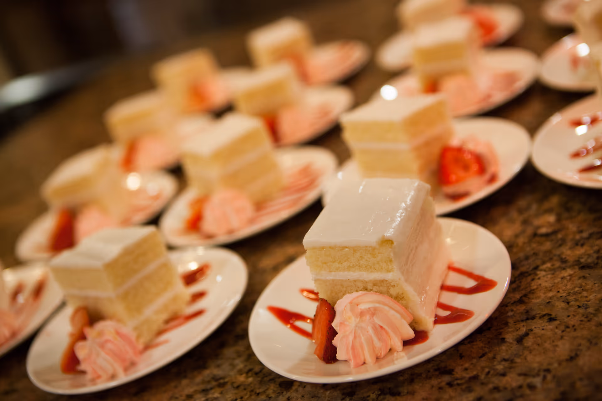 Multiple plates each with a slice of white layered cake topped with white frosting, a dollop of pink whipped cream, a strawberry slice, and a drizzle of red sauce on a granite countertop.
