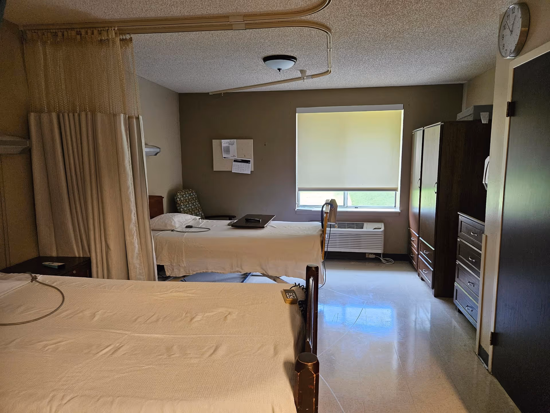 A shared rehabilitation room with two beds separated by a privacy curtain, a window, a chair, and dressers.