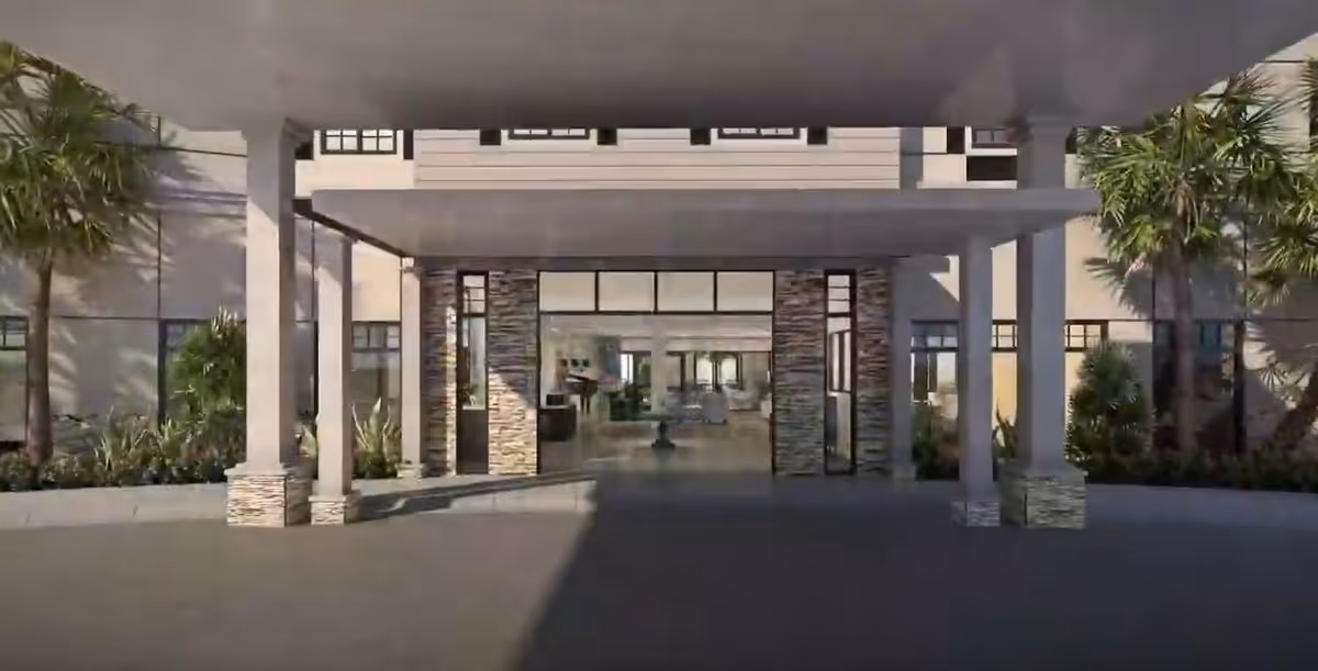 Entrance area of a senior living facility with a covered drop-off zone supported by columns. The building facade features stone accents and large glass doors leading inside. Palm trees and landscaping are visible on either side of the entrance.