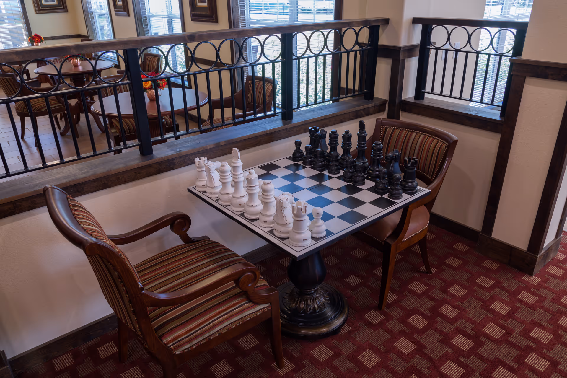 A cozy indoor seating area with a chess table set up for a game, featuring large white and black chess pieces. Two wooden chairs with striped cushions are placed on either side of the table. The area has a red patterned carpet and is adjacent to a railing overlooking a dining area with round tables and chairs near large windows.