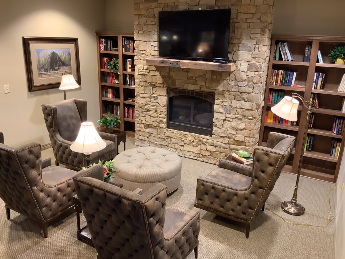 A cozy living room with four tufted armchairs arranged around a round ottoman. The room features a stone fireplace with a mounted flat-screen TV above it. There are two wooden bookshelves filled with books and decorative plants on either side of the fireplace. Several lamps provide warm lighting, and a framed picture hangs on the wall.