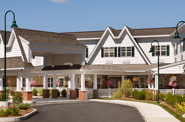 Exterior view of a senior living facility with white siding, multiple peaked roofs, and a covered entrance supported by white columns with brick bases. The building is surrounded by landscaped greenery and hanging flower baskets, with a paved driveway and sidewalk in front.