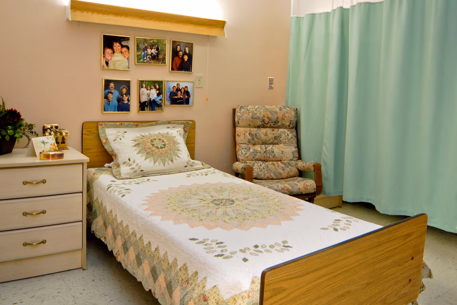 A tidy single bedroom with a wooden bed covered by a floral quilt, a nightstand with decorations, an upholstered armchair, family photos on the wall, and a mint-green privacy curtain.