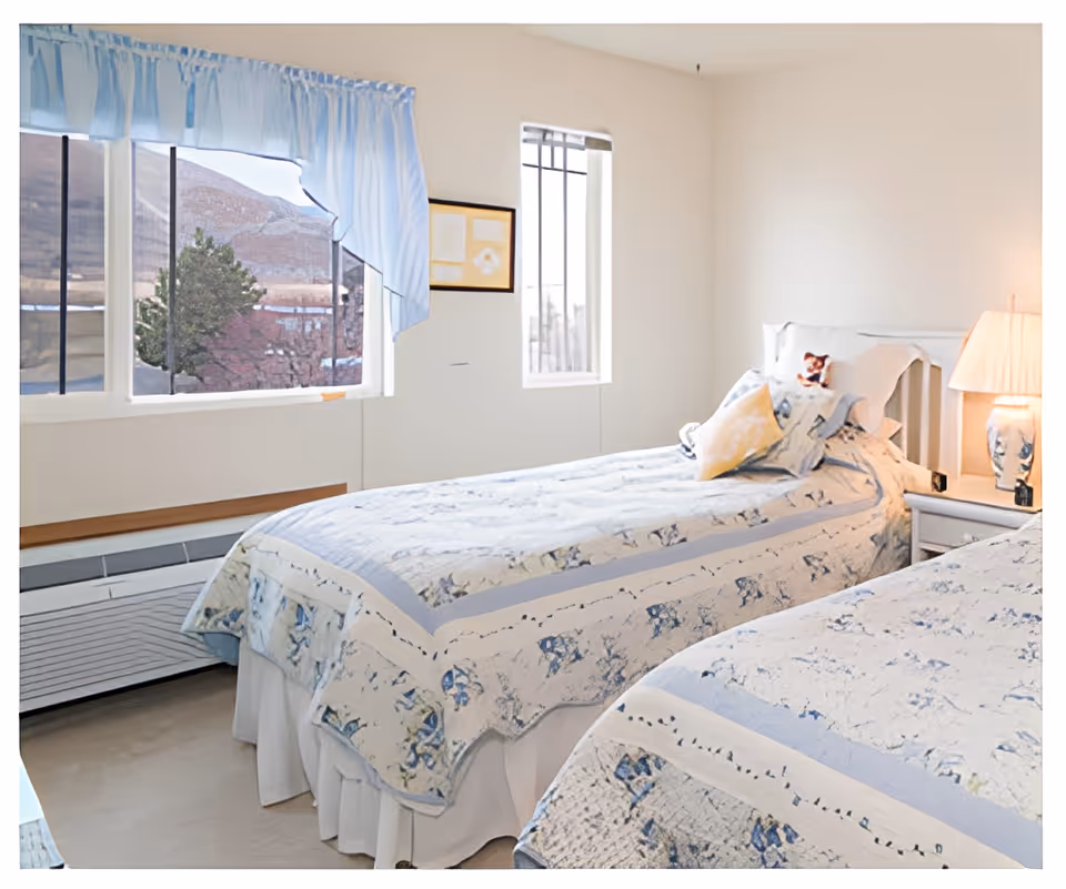 Two twin beds with blue-and-white floral bedspreads in a bright bedroom with a nightstand, lamp, and windows.
