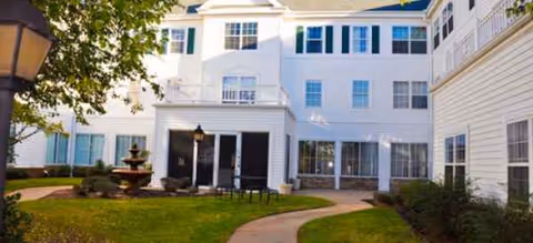 White multi-story senior living building with curved walkway, fountain, and manicured lawn.