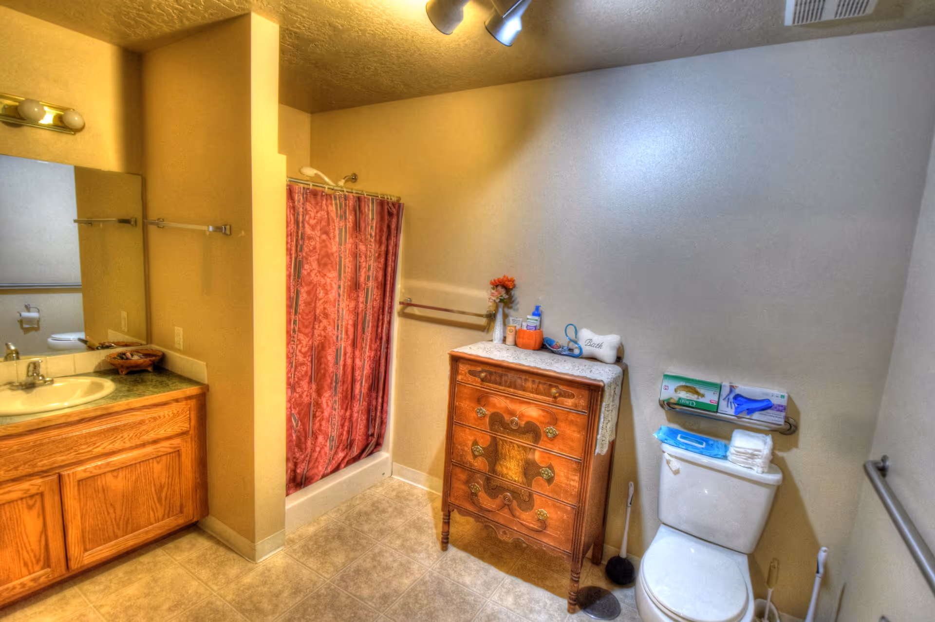 A bathroom with a wooden vanity and sink on the left, a shower with a red patterned curtain in the center, and a toilet on the right. Next to the toilet is a wooden chest of drawers with various toiletries and a small vase with flowers on top. The walls are light-colored, and the floor is tiled.