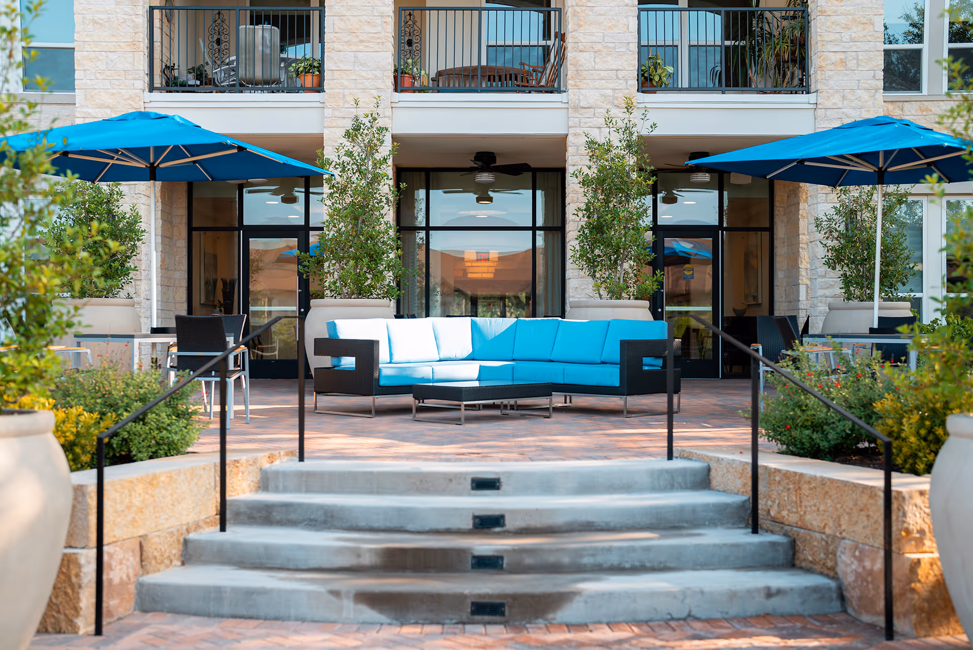 Outdoor patio area with a blue cushioned sectional sofa, two blue umbrellas, potted plants, and steps leading up to glass doors of a building.