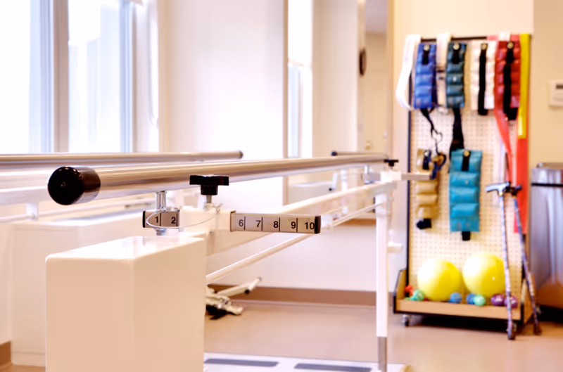 Physical therapy room with parallel bars for walking exercises and a rack holding various weighted straps and exercise balls.